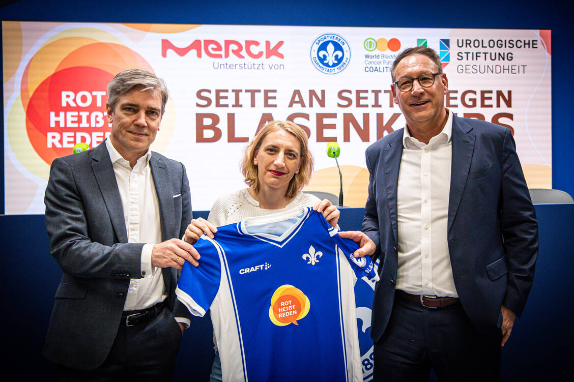 Seite an Seite die Früherkennung von Blasenkrebs bekannter machen: Dr. Matthias Wernicke von Merck (links) und Dr. Severine Banek von der Urologie des Uniklinikums Frankfurt präsentieren zusammen mit Lilien-Präsident Rüdiger Fritsch das Bundesliga-Sondertrikot.  / Foto: SV98